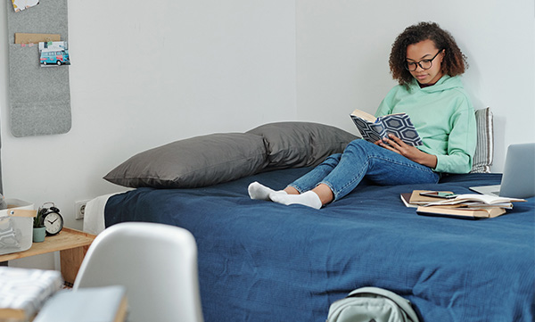person sitting on a bed reading a book in a cozy bedroom with a laptop and books on a nearby table relaxing space for studying and leisure activities