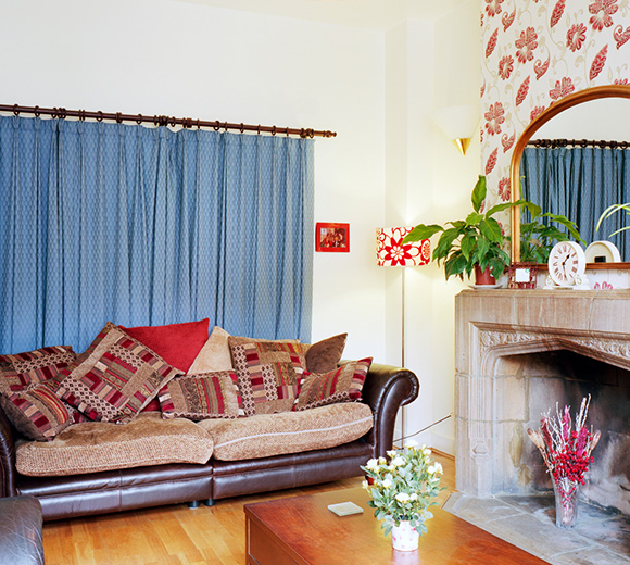 a cozy living room featuring a brown leather sofa adorned with several decorative cushions a blue curtain covers a window and a small coffee table sits in front of the sofa vibrant floral wallpaper adds color to the space a plant and decorative items are present creating a warm and inviting atmosphere suitable for relaxation and gatherings in a home with 6 decorative elements
